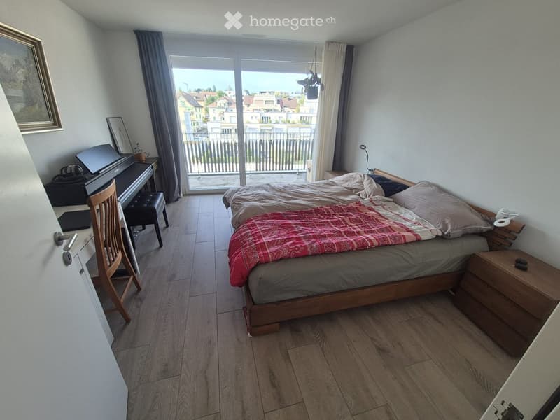 Cozy bedroom with a wooden bed, piano, chair, and nightstand. Large window with balcony access offers a view of the city.
