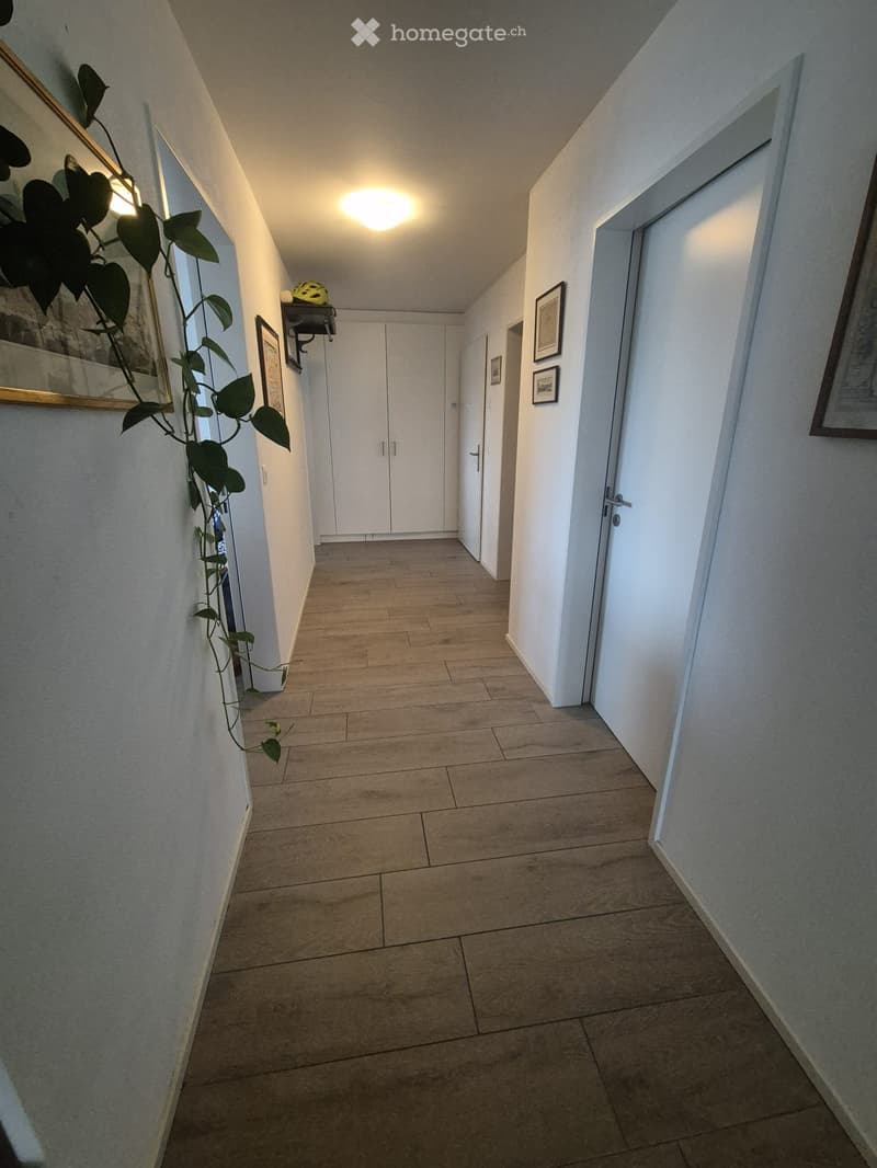 Clean hallway with tiled floor, white walls, and several doors leading to rooms. Features framed artwork and a small plant on the left side.