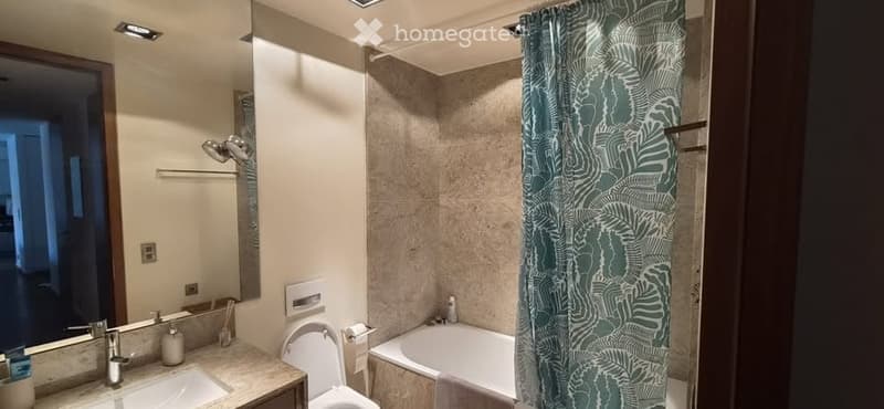 Modern bathroom with a white bathtub, toilet, sink, and a large mirror. The shower area is separated by a green leafy-patterned curtain. The walls are tiled in a neutral tone, and the space is well-lit with recessed ceiling lights.
