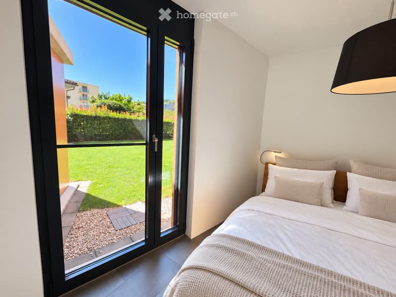 Modern bedroom with a large bed, white bedding, and a view of a well-maintained garden through large glass doors.