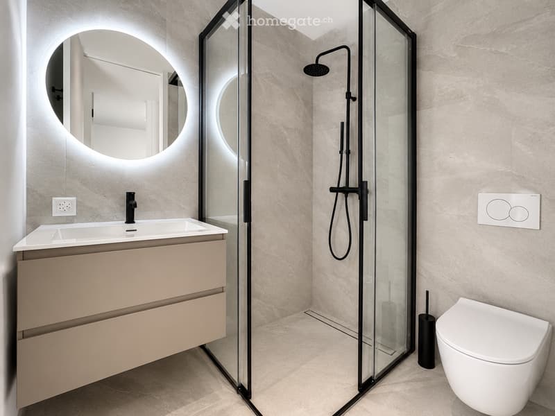 Modern bathroom with beige tiled walls and floor, a floating vanity with a sink and faucet, a round illuminated mirror, a glass-enclosed shower with a handheld showerhead, and a toilet.