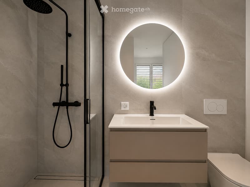 Modern bathroom with a white sink, beige vanity, round illuminated mirror, black showerhead, and a toilet.