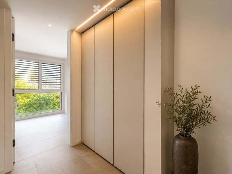 Modern hallway with white walls, recessed lighting, large window with blinds, and a tall decorative vase with greenery on the right side.