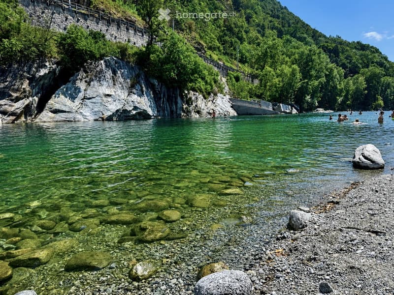 A serene river surrounded by lush green trees and rocky cliffs, with people enjoying the water and a stone bridge in the background.