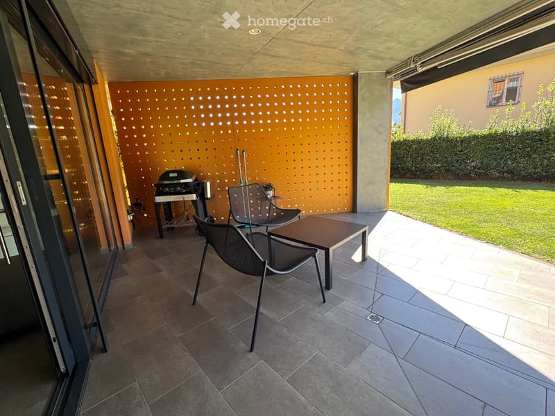 Balcony with tiled floor, modern furniture including a table and chairs, and a grill against an orange perforated wall. The balcony has large glass doors leading inside and overlooks a well-maintained lawn and garden area.