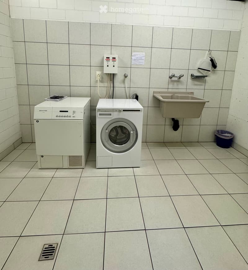 A clean, tiled bathroom with a washing machine, dishwasher, sink, and a small trash bin.