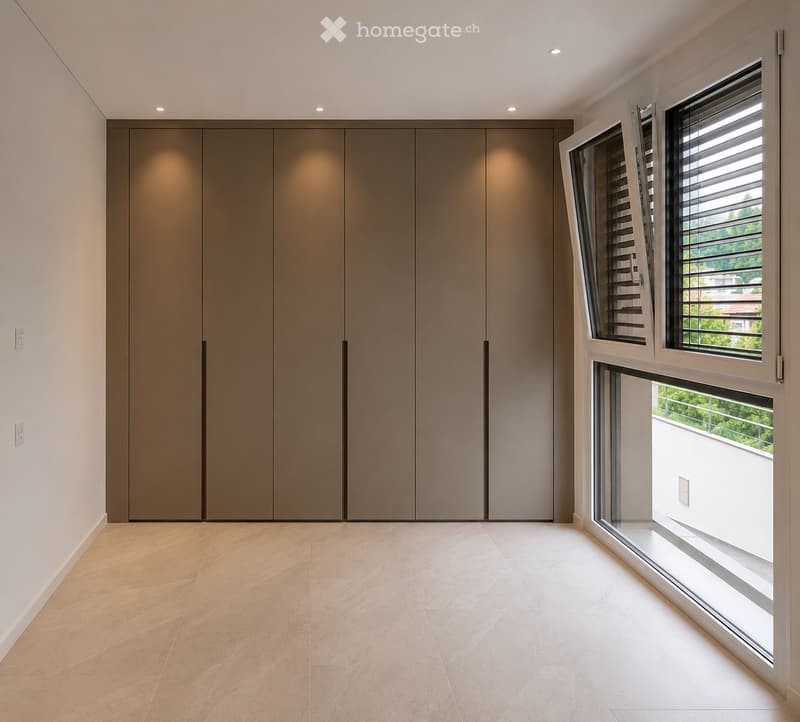 Empty room with beige tiled floor, large built-in wardrobe, and large windows with blinds, offering a view of greenery outside.