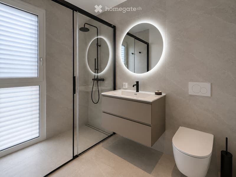 Modern bathroom with a sleek sink, round illuminated mirror, toilet, and a glass-enclosed shower area.