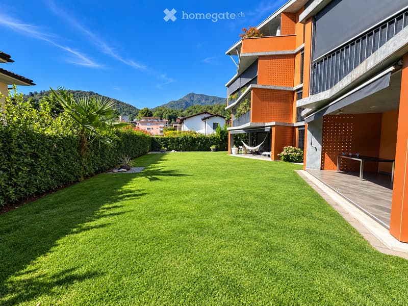 Well-maintained lawn with trimmed hedges, a modern building with orange accents, and a hammock on the patio.