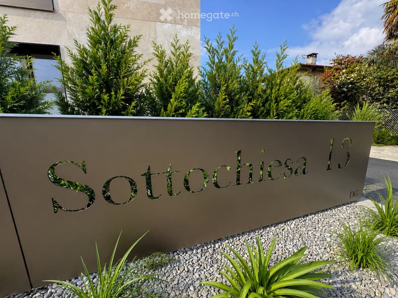 A modern outdoor area with a sleek sign reading "Sottochiesa 13" on a stone wall, surrounded by neatly trimmed shrubs and a gravel pathway.