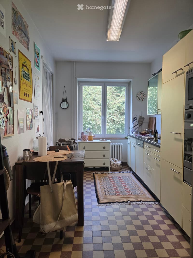 Bright kitchen with checkered floor, white cabinets, and a large window overlooking greenery. Includes a wooden dining table with chairs, a small cabinet, and various kitchen items.
