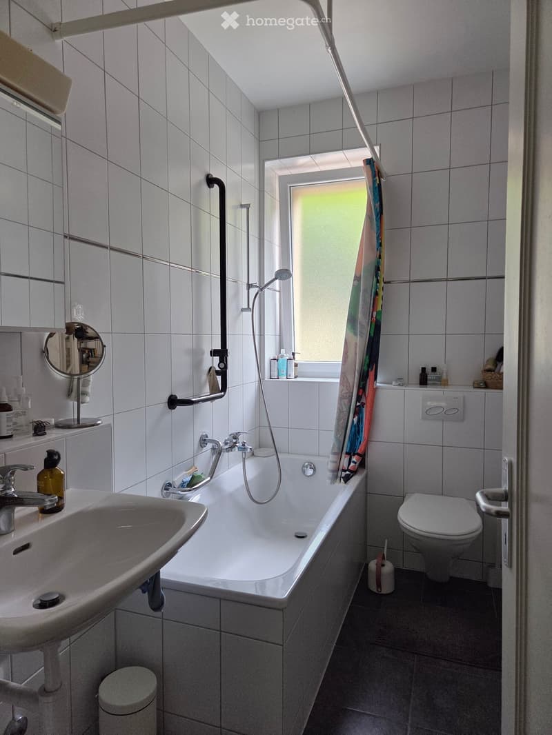 Bathroom with white tiled walls, bathtub, toilet, sink, mirror, and shower area with a curtain. Includes a small window and various toiletries on the windowsill.