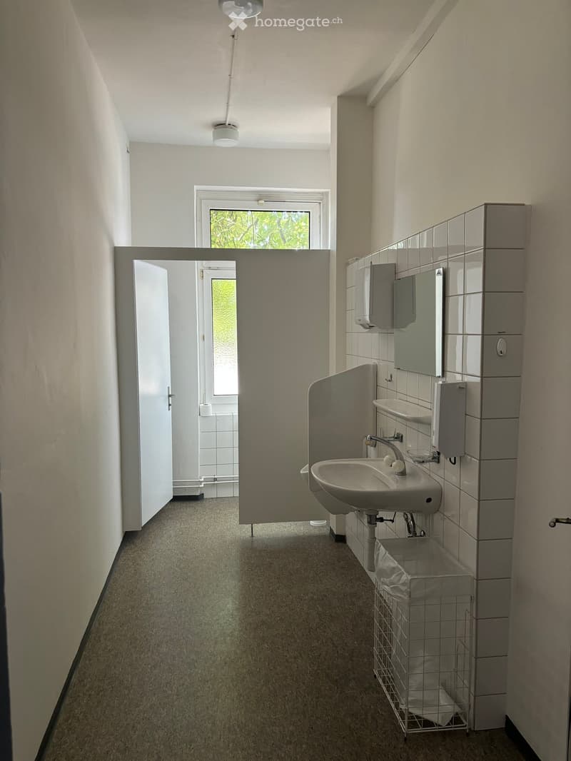 Clean, well-lit hallway with carpeted floor, white walls, and a window at the far end. Features a sink area with a mirror, soap dispenser, and a trash bin.