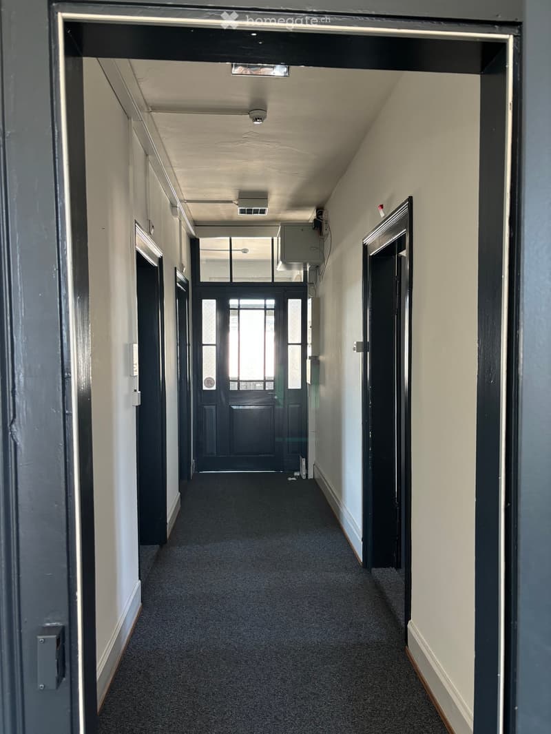 Empty hallway with beige walls, dark carpet, and several doors, including a central black door with glass panels.