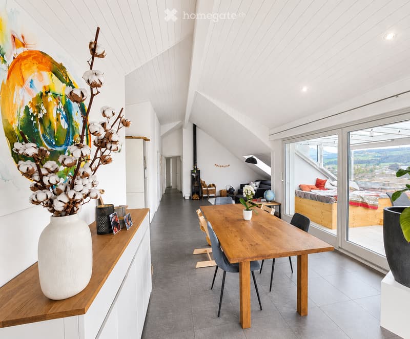 Modern, open-plan living and dining area with wooden table, chairs, white vase with cotton flower arrangement, picture frames, sofa, bed, globe, and large windows offering a scenic view.