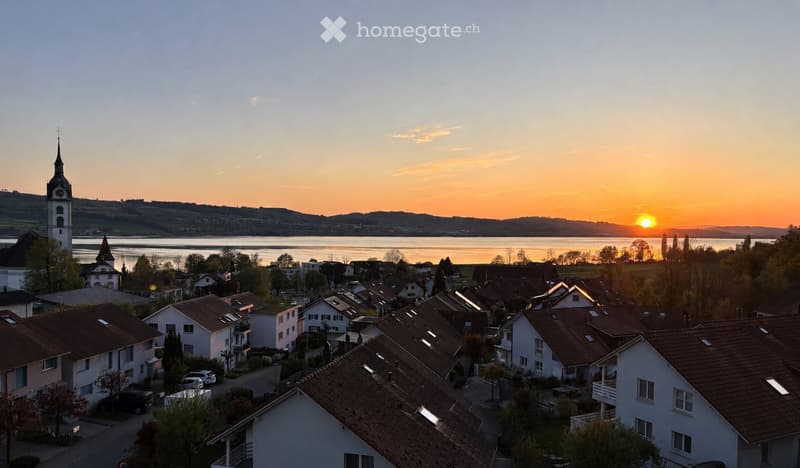 A village scene at sunset with houses, a church tower, and a body of water in the background.