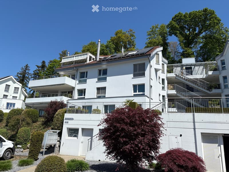 A modern, multi-story white house with balconies, large windows, and a staircase leading to the entrance. The house is surrounded by greenery and trees.