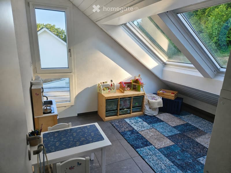 A cozy, well-lit attic room with skylights, featuring a small kitchenette, a play table, a toy box, and a colorful rug.