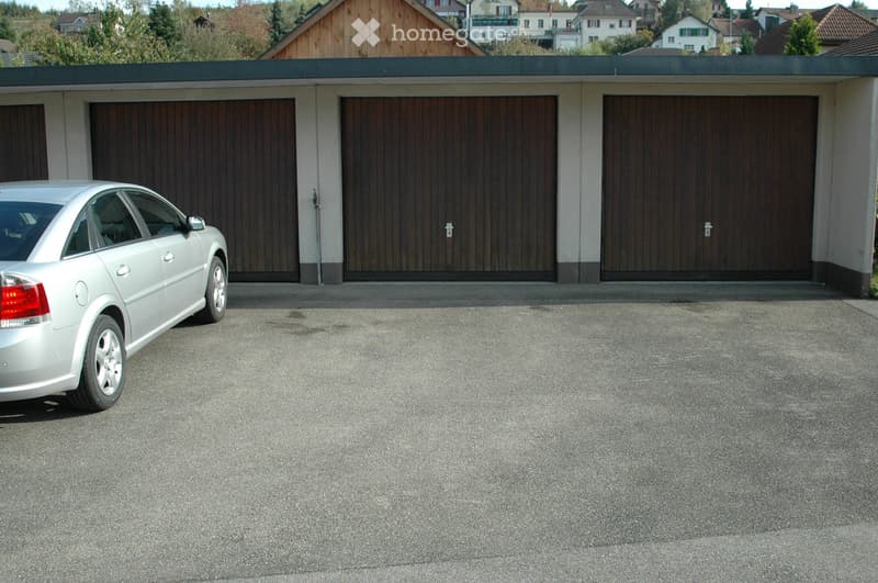 A modern garage with three large wooden doors, a silver car parked in front, and a smooth concrete driveway.