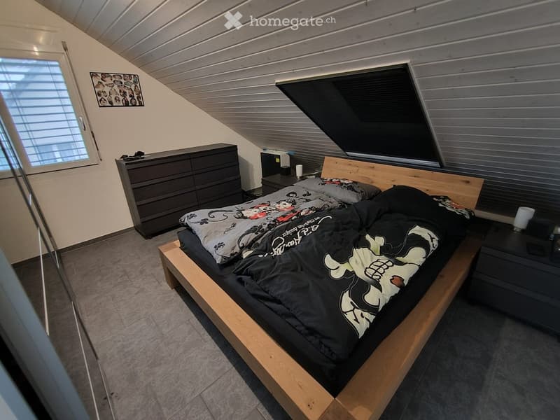 Modern bedroom with a wooden bed frame, black bedding, nightstands, and a dresser. The room features slanted ceilings and large windows.