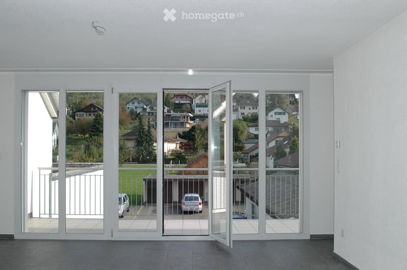 Bright, modern hallway with large, open sliding glass doors leading to a balcony and outdoor view of a residential area with houses, trees, and parked cars.