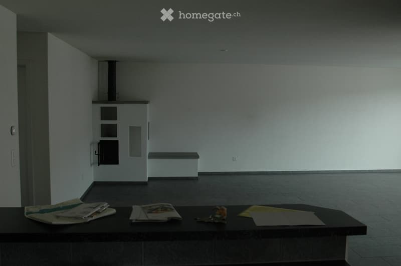 A small, unfurnished room with a dark wooden table holding magazines, a toy, and some papers. The room has white walls, a tiled floor, and a built-in bench along one wall.