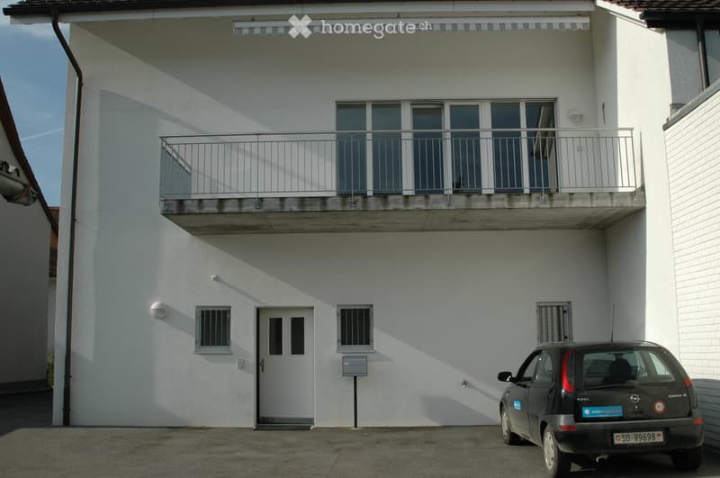 Two-story white building with a balcony on the upper floor, a covered entrance, and a parked car in front.