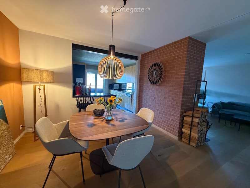Modern dining area with a round wooden table, four chairs, a vase with sunflowers, and a bowl. Adjacent living area features a sofa, a table lamp, and a decorative wooden stack. Exposed brick wall and pendant light visible.