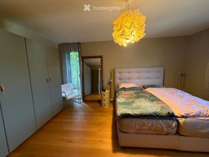 Well-lit bedroom with wooden flooring, a large upholstered headboard bed, a white chair, a mirror, and a decorative hanging lamp.