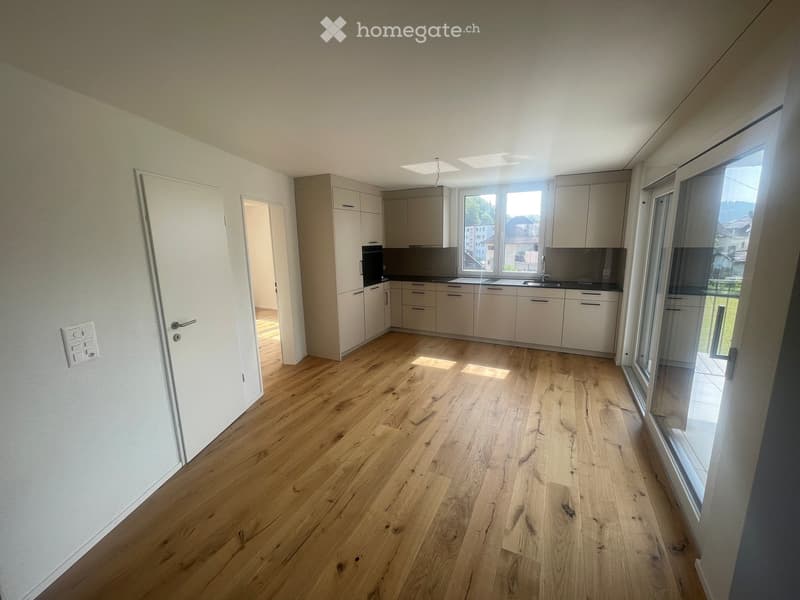 Empty kitchen with light wooden flooring, white cabinets, and large windows offering a view of the outside. No furniture or appliances are present.