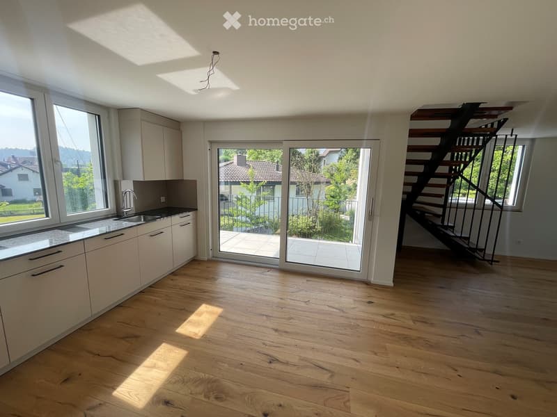 Empty room with light wooden flooring, white cabinets, a sink, and large windows with a view of the garden. A staircase leads to an upper level.