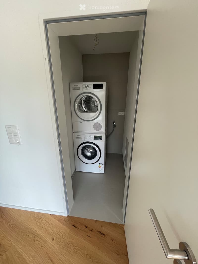 A narrow hallway leads to a compact laundry area with stacked washing and drying machines, light-colored walls, and wooden flooring.