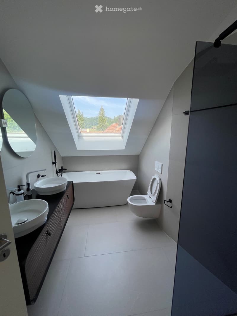 Modern bathroom with a large skylight, white bathtub, two sinks, toilet, and a round mirror.