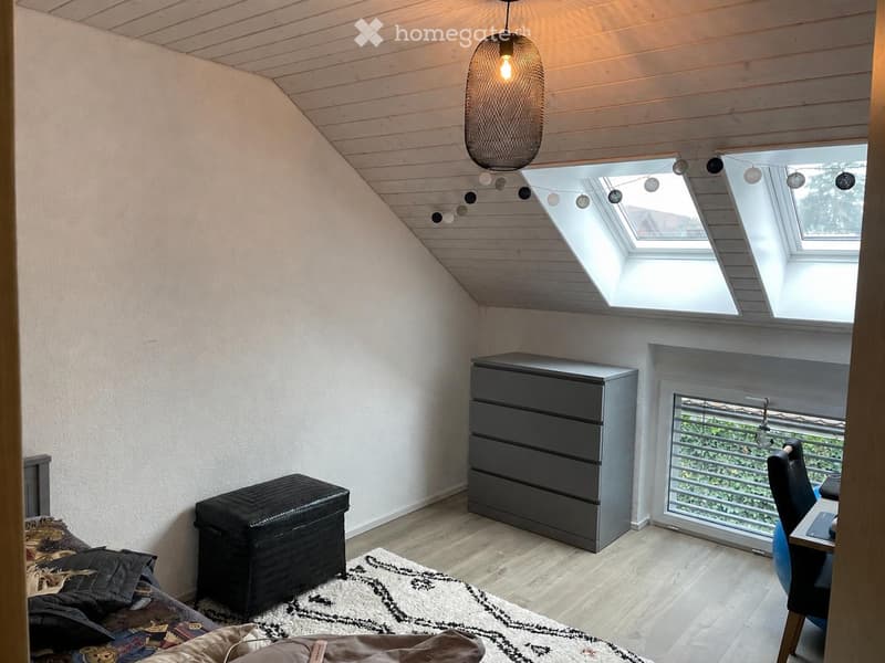 Cozy attic bedroom with wooden slanted ceiling, skylight windows, and modern furniture including a bed, chair, table, chest of drawers, and a stool.