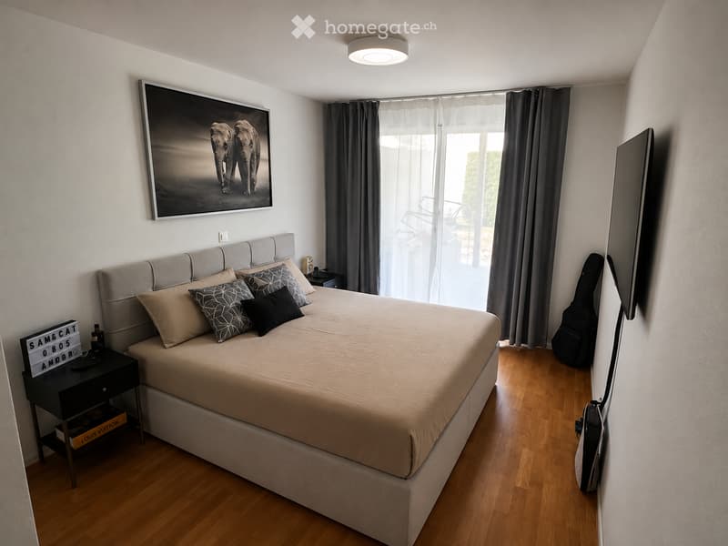 Modern bedroom with a large bed, nightstand, picture frame, and guitar case. The room features wooden flooring and large windows with curtains.