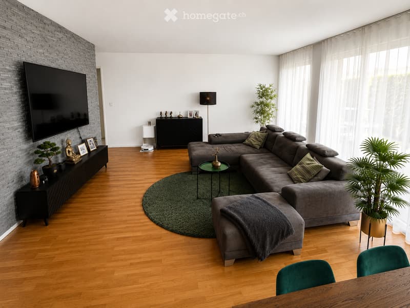 Modern living room with hardwood flooring, grey sectional sofa, coffee table, ottoman, side tables, television, and decorative plants.