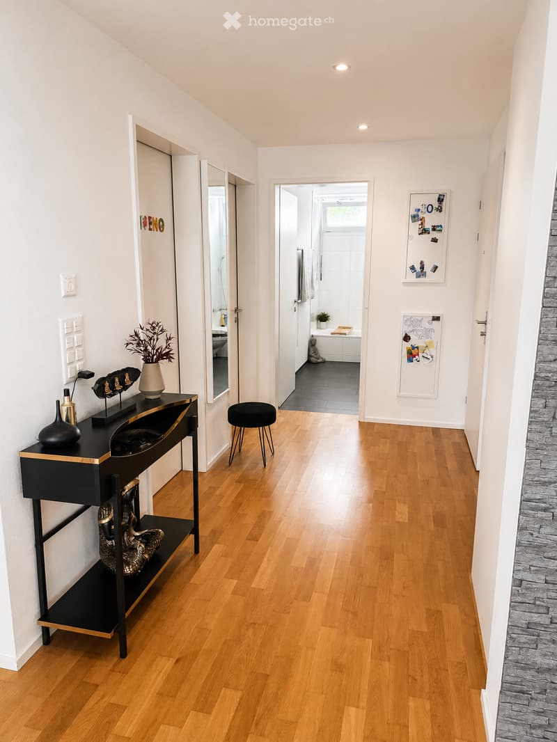Modern hallway with wooden flooring, white walls, and recessed lighting. Features a black console table with decorative items, a small black stool, and a view into a bathroom with a bathtub and white tiles.
