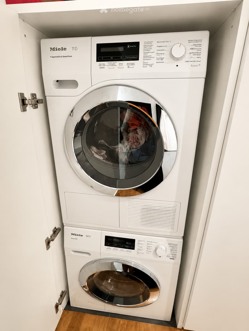 White Miele washing machine and dryer unit installed in a cabinet, with wooden flooring visible.