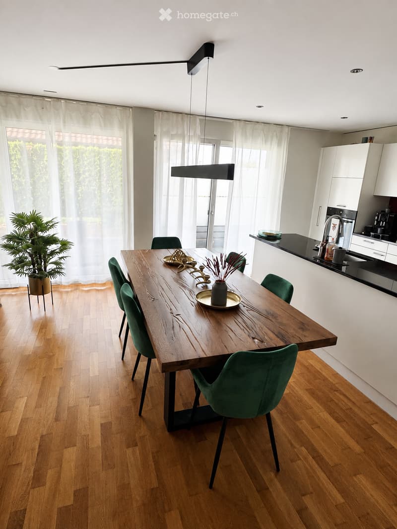 Modern dining area with a large wooden dining table, green chairs, and a sleek kitchen area featuring white cabinets and a black countertop.