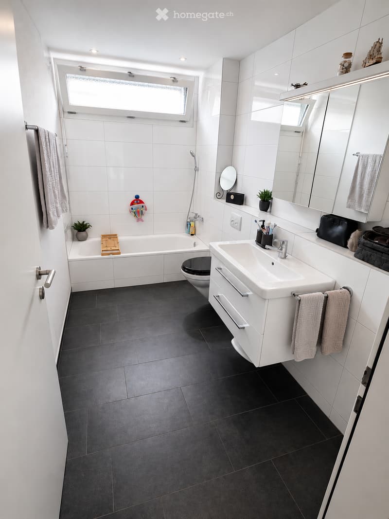 Modern bathroom with white tiles, dark grey floor, bathtub, toilet, sink, mirror, and towel racks.