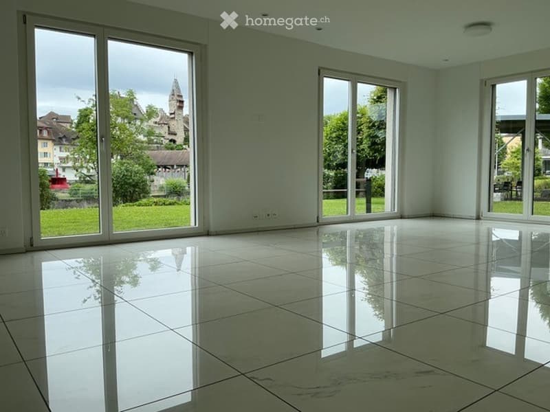 Empty room with large windows, polished tile floor, and a view of a green area and buildings outside.