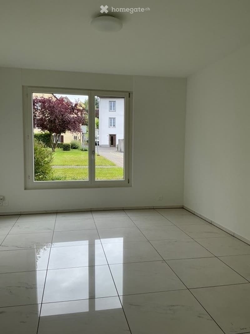 Empty room with tiled floor, large window showing a view of a garden and neighboring house, white walls, and a ceiling light fixture.