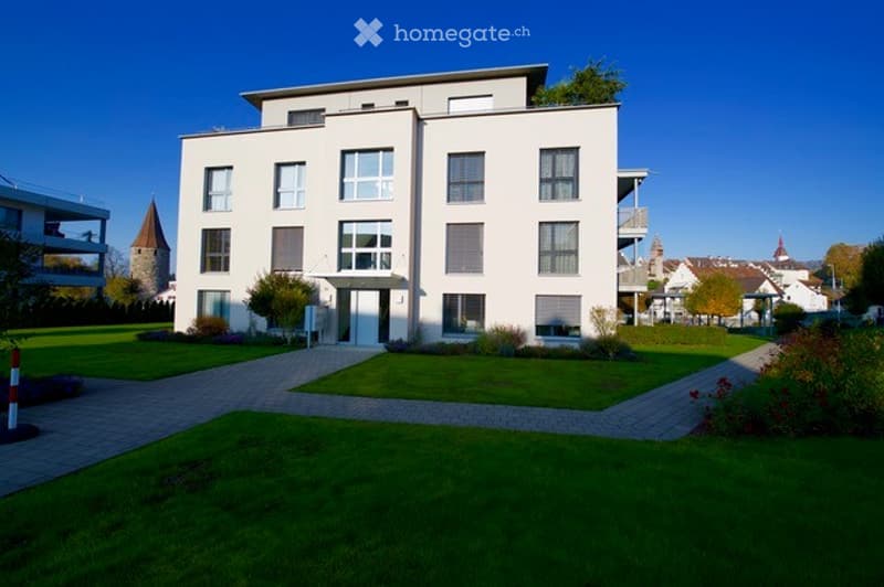 Modern white apartment building with multiple windows and balconies, surrounded by a well-maintained lawn and pathways.