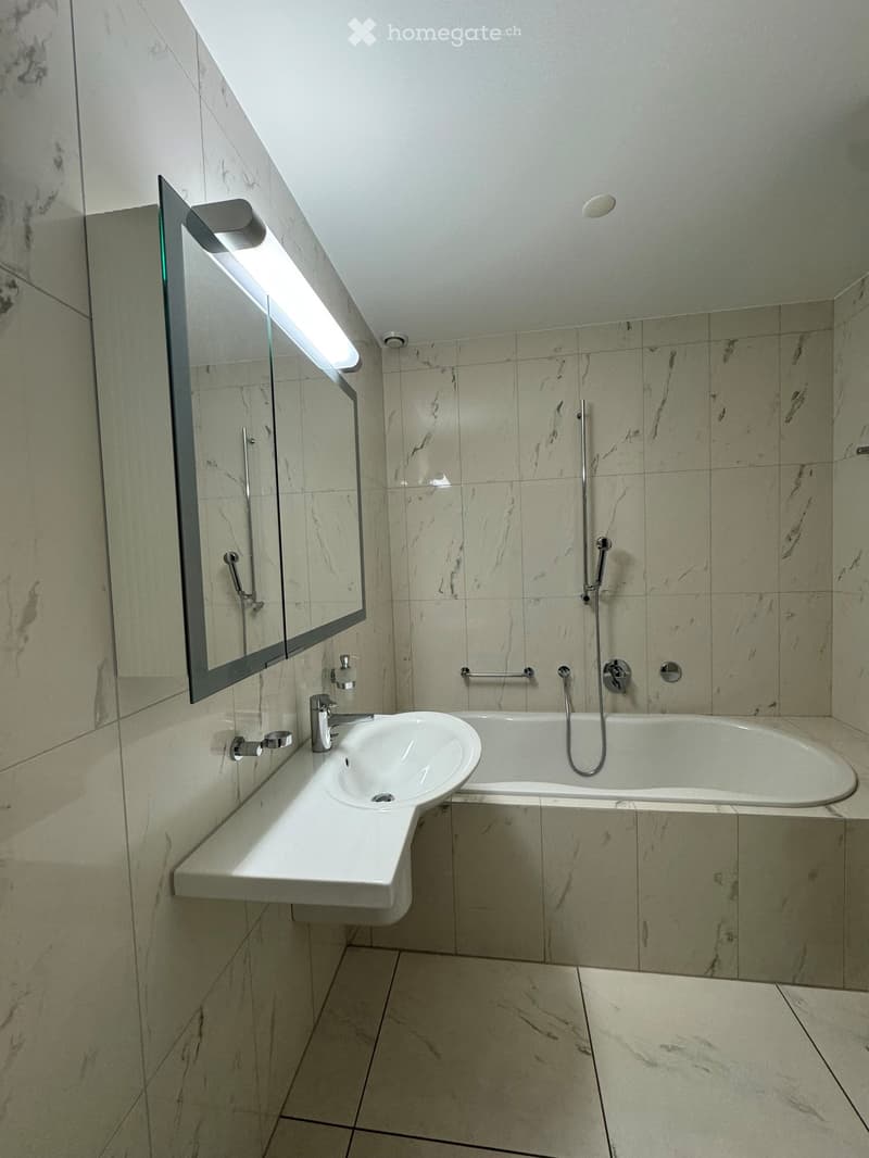 Modern bathroom with marble tiles, a white sink, bathtub, and a large mirror with built-in lighting.