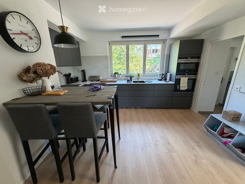 Modern kitchen with grey cabinets, wooden floor, central island, dining table with chairs, and various appliances including an oven, sink, and coffee maker.