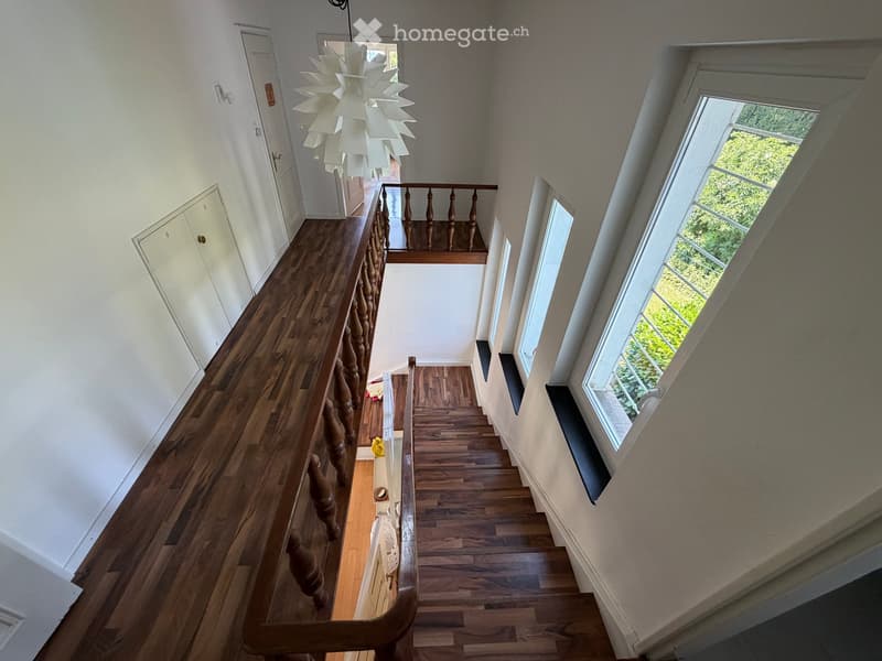Spacious hallway with wooden flooring, white walls, and a staircase leading to an upper level. Features a decorative chandelier and large windows allowing natural light to enter.