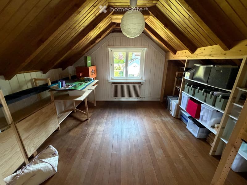 Spacious attic room with wooden floors, slanted wooden ceilings, large window, shelves, and storage boxes.