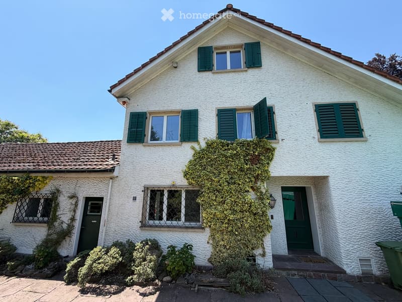 Two-story white house with green shutters, ivy-covered walls, and a tiled roof. Features include a front door, multiple windows, and a small garden area with shrubs.
