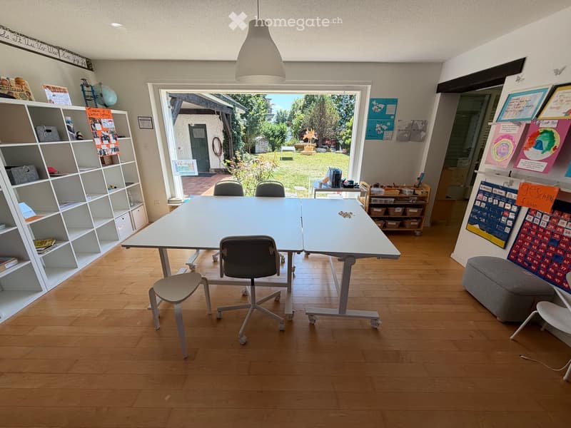 Bright room with wooden flooring, large table, chairs, shelves filled with books and supplies, and a view of a garden through large windows.