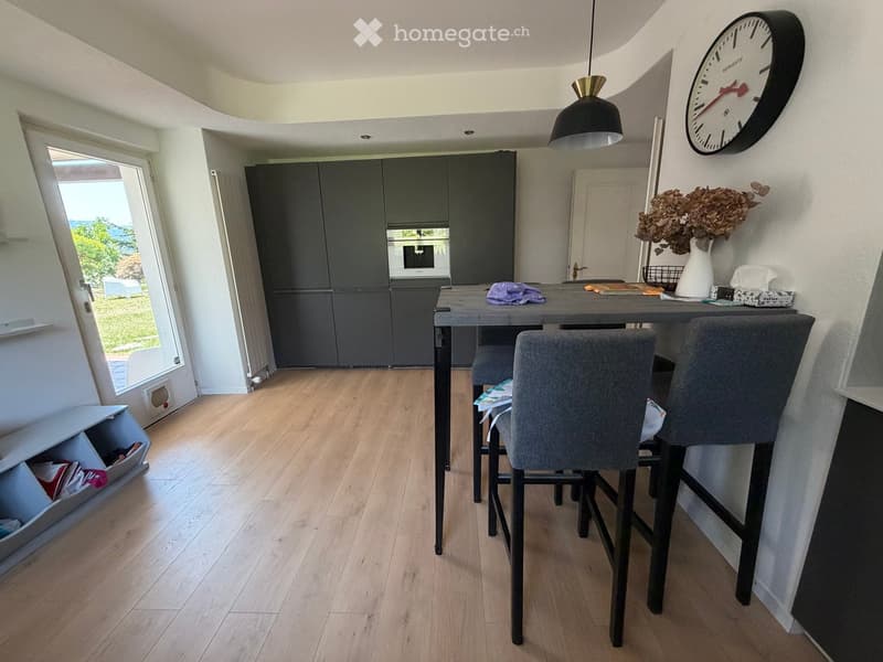 Modern kitchen with grey cabinets, a central island table with two chairs, a vase with dried flowers, and a wall-mounted clock.